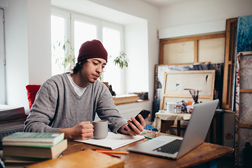 Man at a desk drinking coffee, looking at his phone, with his laptop open.